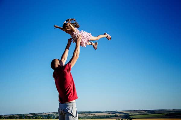 A man in a red shirt joyfully lifts a young child wearing a polka dot dress high into the air against a bright blue sky, capturing a moment of trust, happiness, and playfulness.