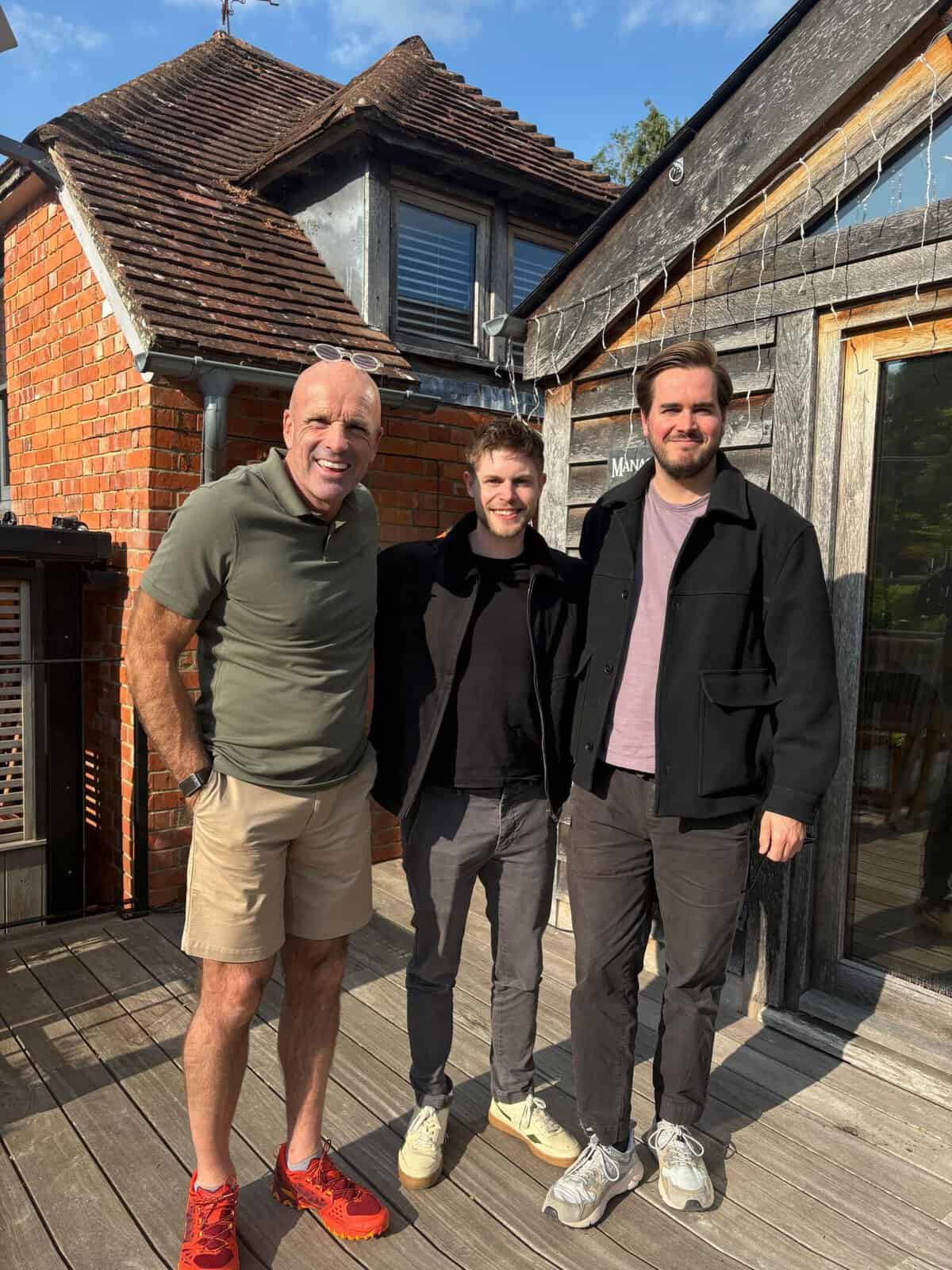 Three men standing outdoors in front of a rustic building with wooden and brick elements.