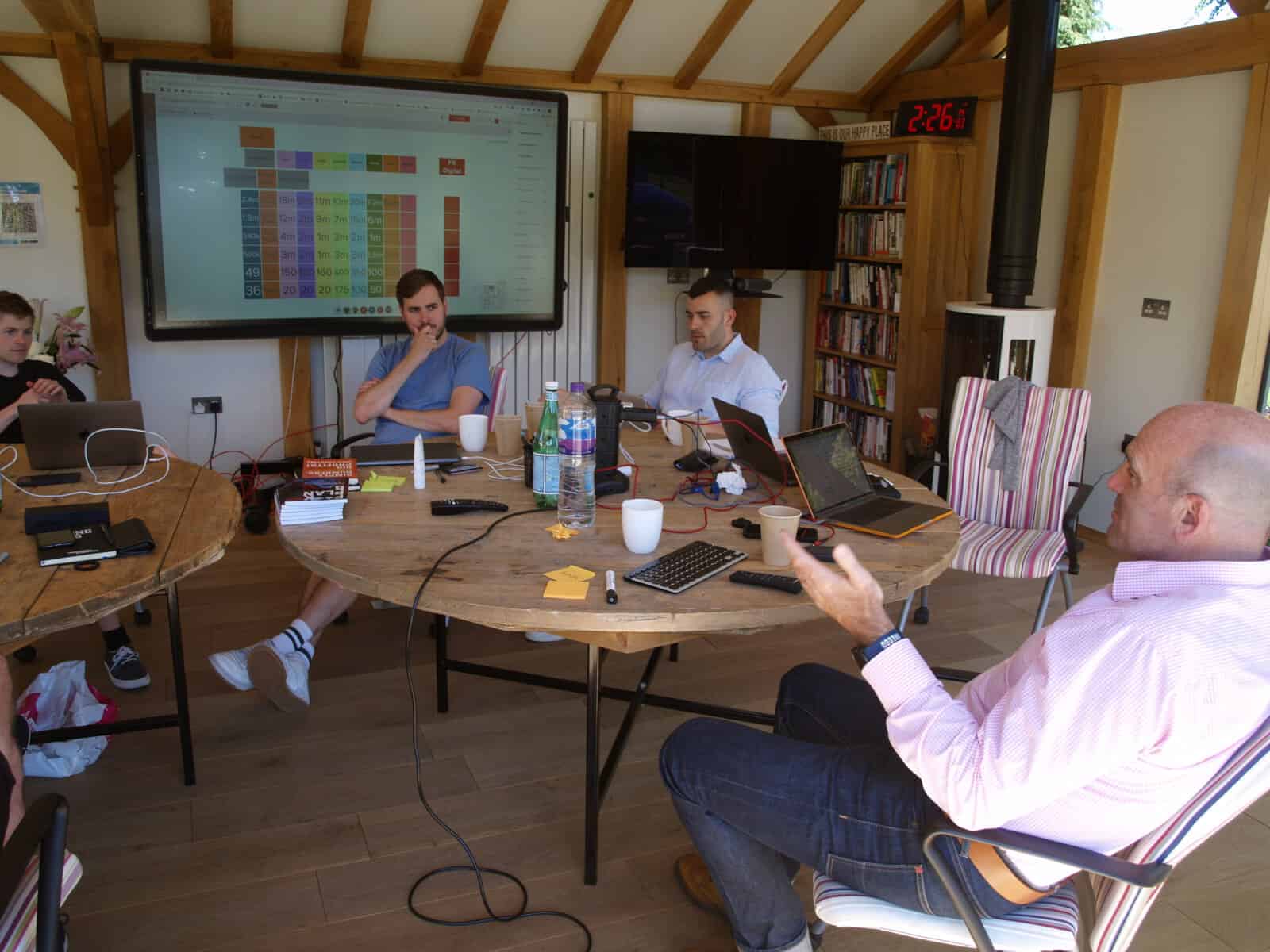 Four team members in a daily huddle meeting around a table with a data chart on screen