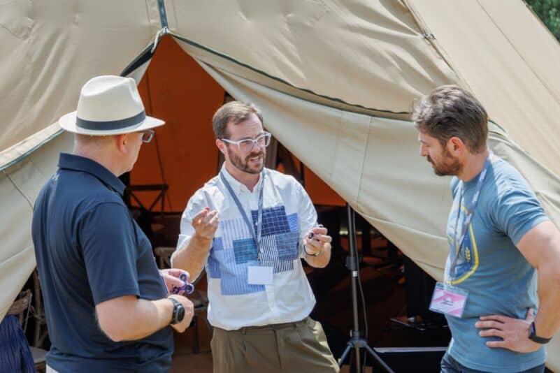 Three men engaging in conversation at an outdoor event with a tent in the background.